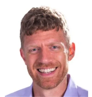 A smiling man with curly light brown hair and a beard, wearing a light blue shirt.