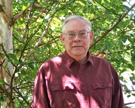Older man with glasses and white hair wearing a maroon shirt, standing outdoors against a leafy green background.