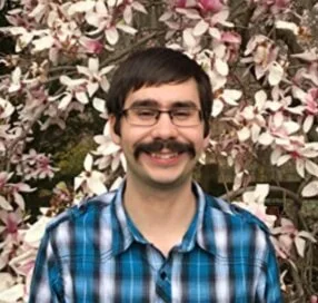 A man with glasses and a mustache smiling, wearing a blue plaid shirt, standing in front of pink and white flowers.