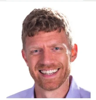 A smiling man with curly brown hair and a beard, wearing a light blue shirt, against a plain white background.