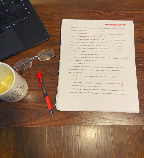 A wooden desk with a black laptop, a pair of reading glasses, a red and black pen, a cup with yellow liquid, and a stack of printed papers with handwritten notes and red markings.