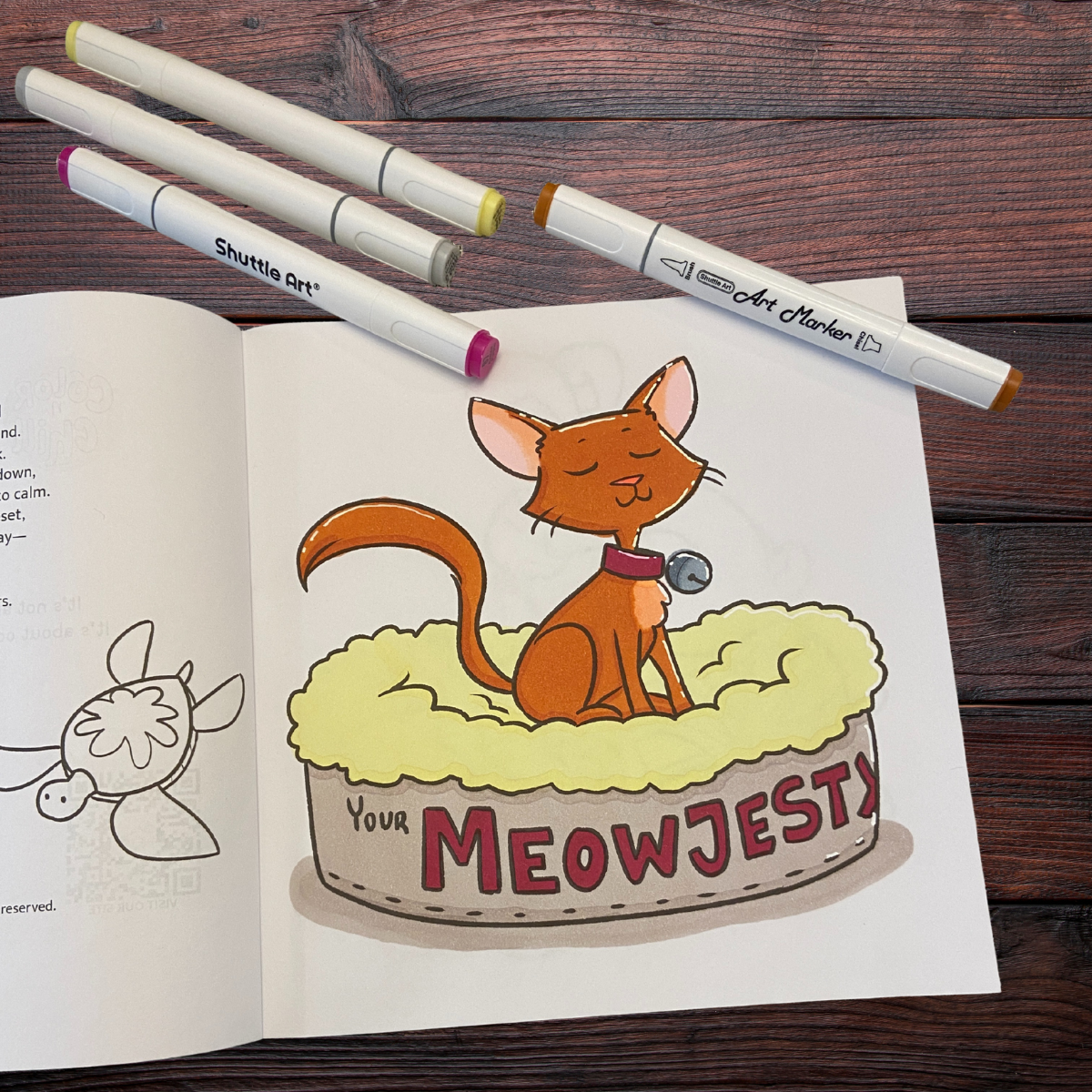 Colorful illustration of a happy, content brown cat with a collar and a bell, sitting in a plush yellow bed with the words "Your MEOWJESTY" on the side, surrounded by drawing markers on a wooden surface.