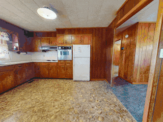 Empty kitchen with wooden cabinets, white refrigerator, and basic appliances in a room with wood-paneled walls and a linoleum floor.