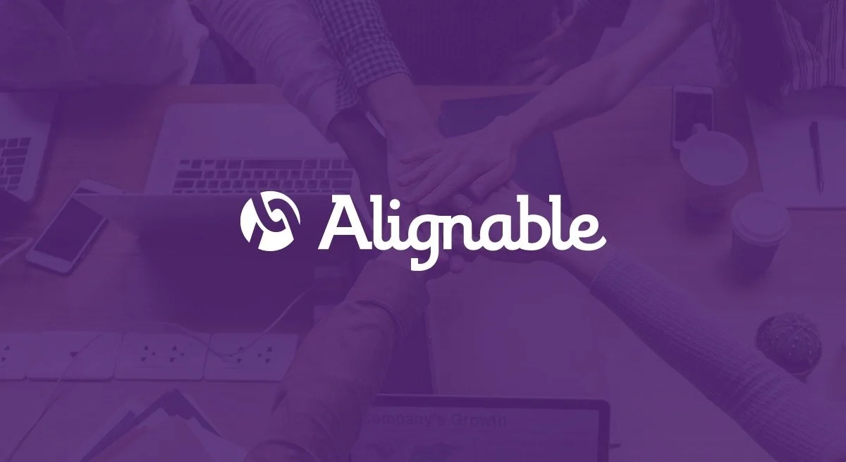 People doing a team fist bump in an office setting with laptops and coffee cups on the table, overlaid with the Alignite logo.
