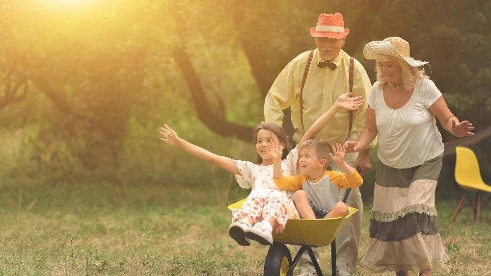 Two children riding in a yellow wheelbarrow, being pushed by an older man and woman in a sunlit outdoor setting.
