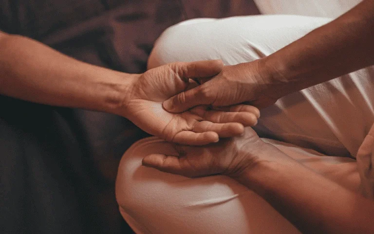 Two people holding hands gently, one sitting with crossed legs wearing light-colored pants, the other resting their hand on the person's knee.