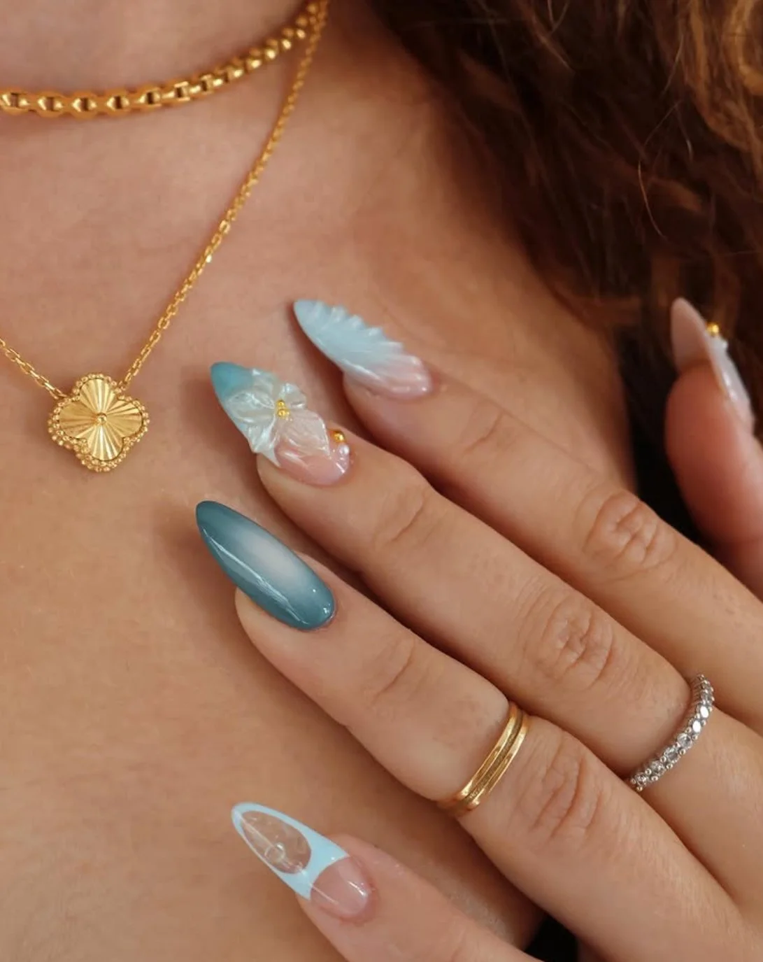 Close-up of a woman's hand with manicured nails resting on her chest. She is wearing layered gold rings, two necklaces including one with a gold flower pendant, and has nail art in shades of blue and white with iridescent accents.