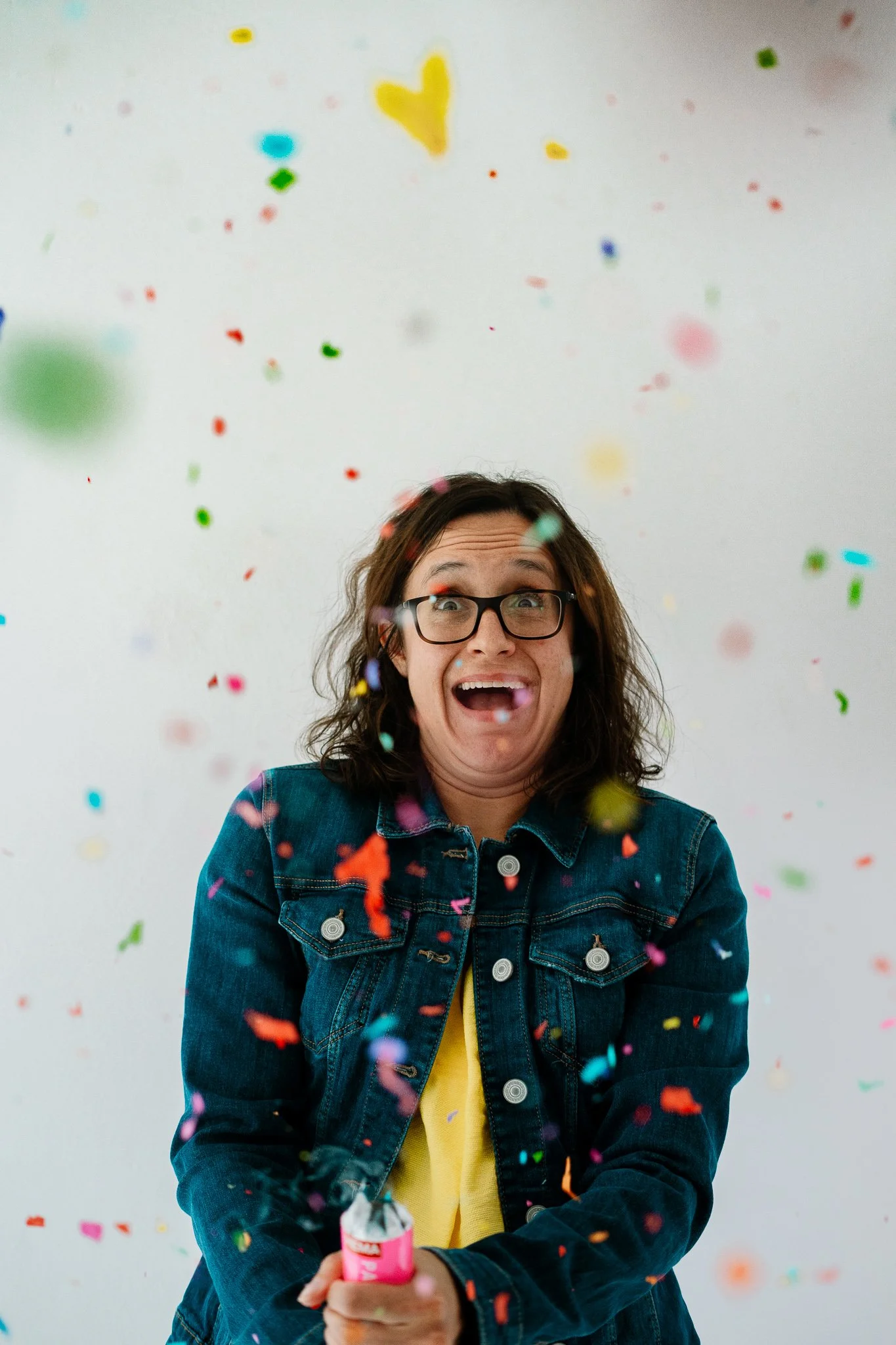 Een vrouw in een denim jas en een gele top die een confettikanon vasthoudt terwijl gekleurd confetti in de lucht vliegt, met een witte achtergrond.