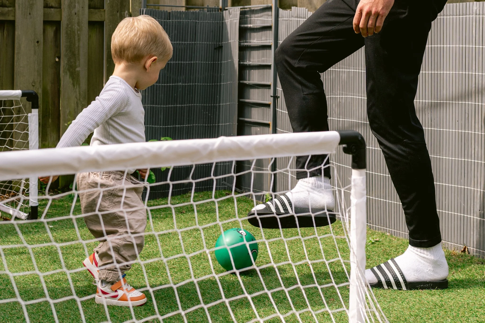 Een kind en een volwassene die samen buiten op gras spelen met een groene voetbal, achter een klein hek. De volwassene is gekleed in zwarte sportkleding en de jongen draagt een witte trui en beige broek.