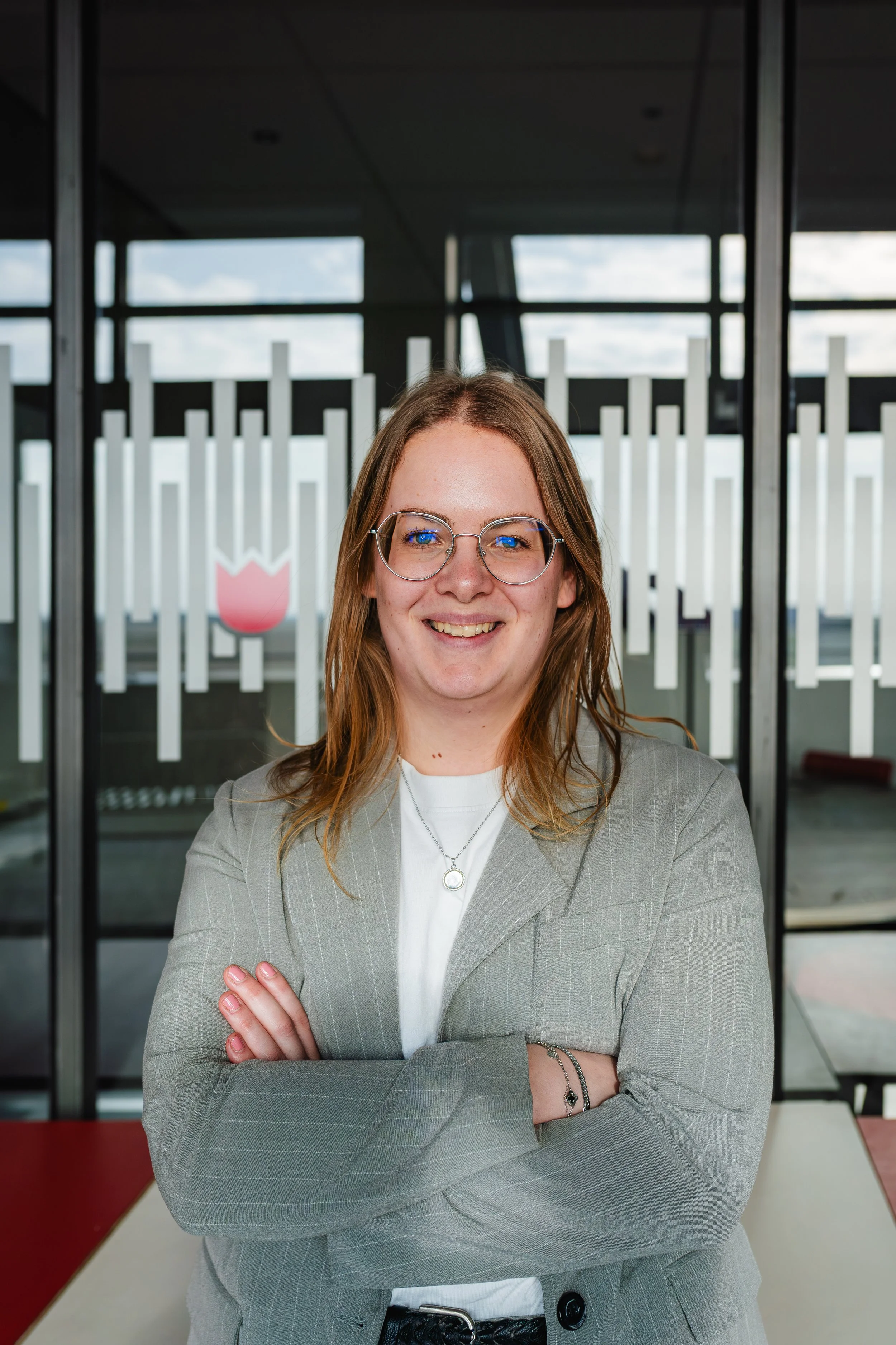 A woman with shoulder-length light brown hair, glasses, and a light gray pinstripe blazer standing with arms crossed, smiling in front of a window with a geometric design, in an indoor setting.