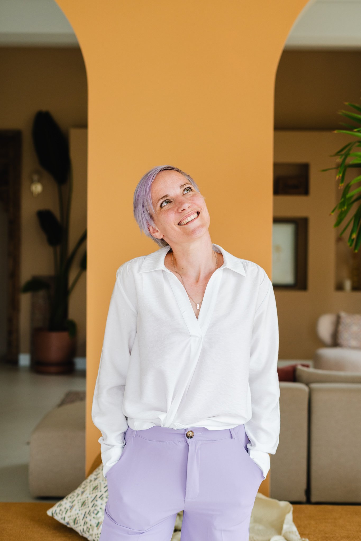 Vrouw met kort, lila haar die glimlacht en naar boven kijkt, in een witte blouse en lichtpaarse broek, binnen in een woning met een gele muur. Een kleurrijke branding fotoshoot van een vrouwelijke ondernemer die andere creatieve ondernemers coacht