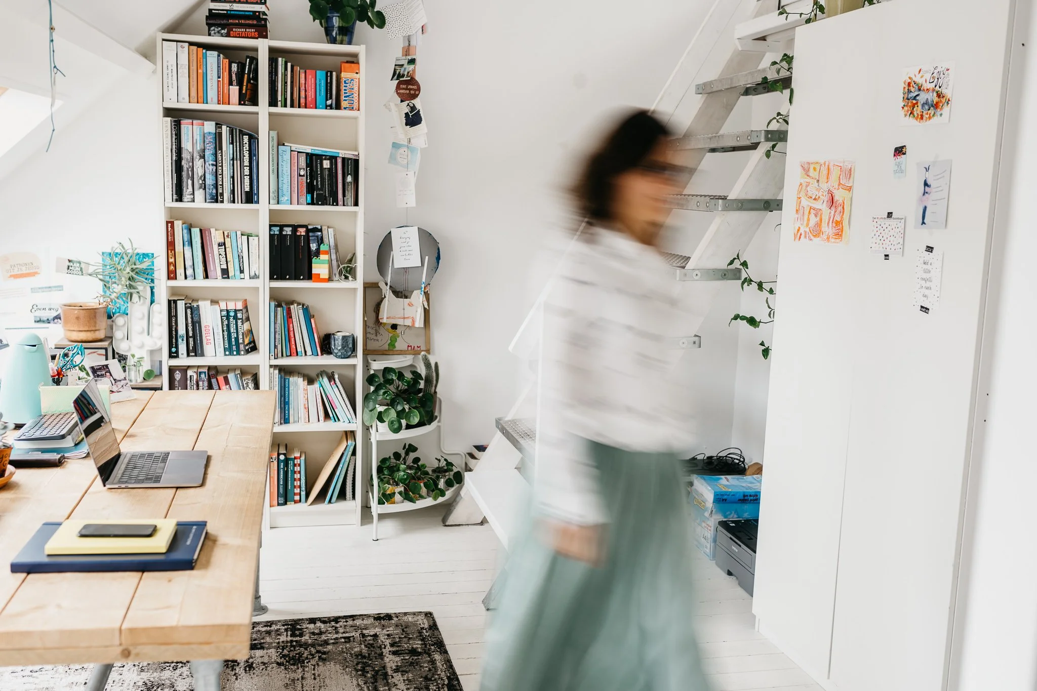 Een overzicht van een huiselijke werk- of leesruimte met een boekenkast vol boeken, een tafel met laptops, pennen en notitieboeken, en een trap die omhoog leidt. Er hangen kleurrijke tekeningen en kaarten aan de muur en er staan diverse planten.