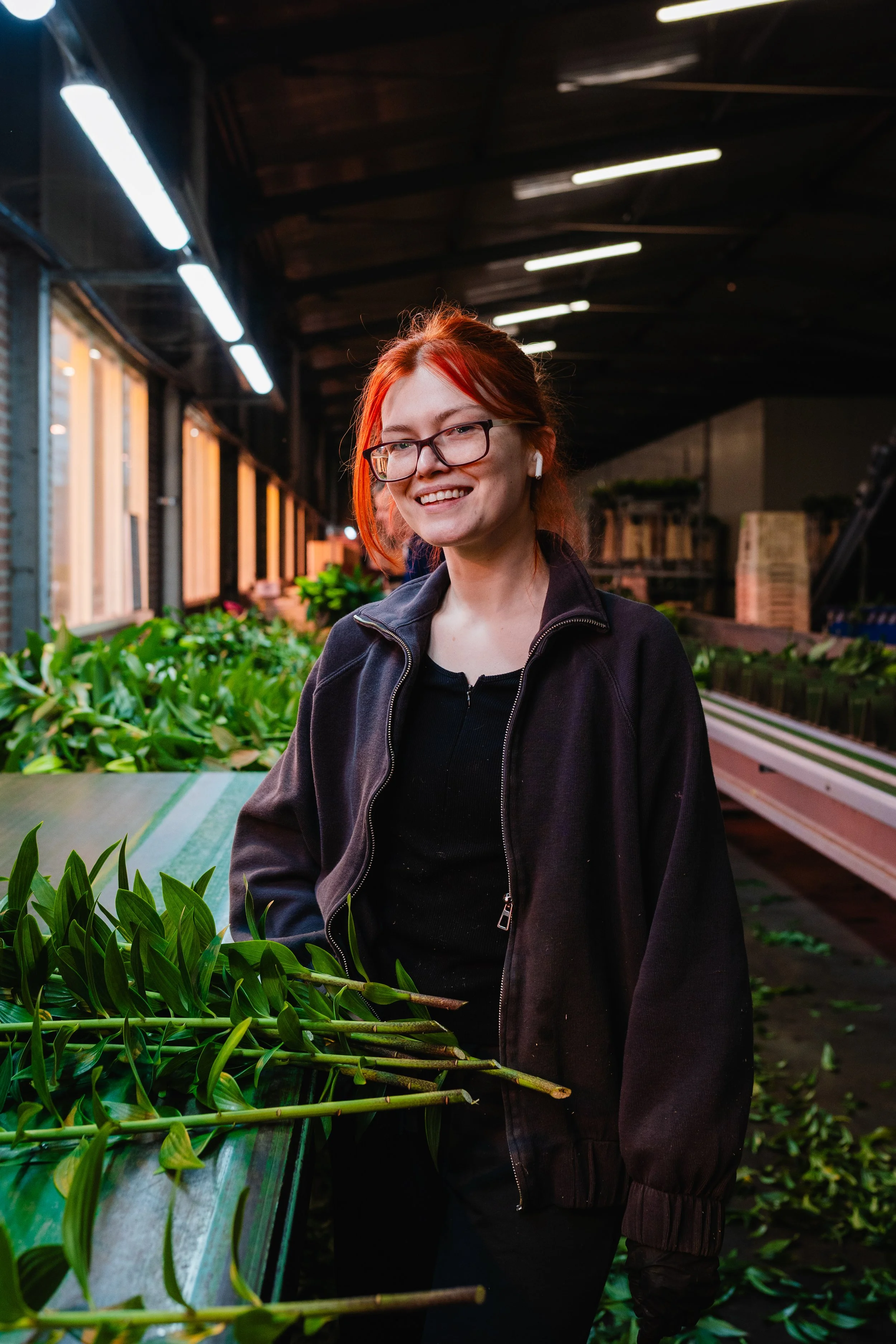 A young woman with red hair, glasses, and wireless earbuds standing inside a plant nursery with green leafy plants on a table and large windows behind her.