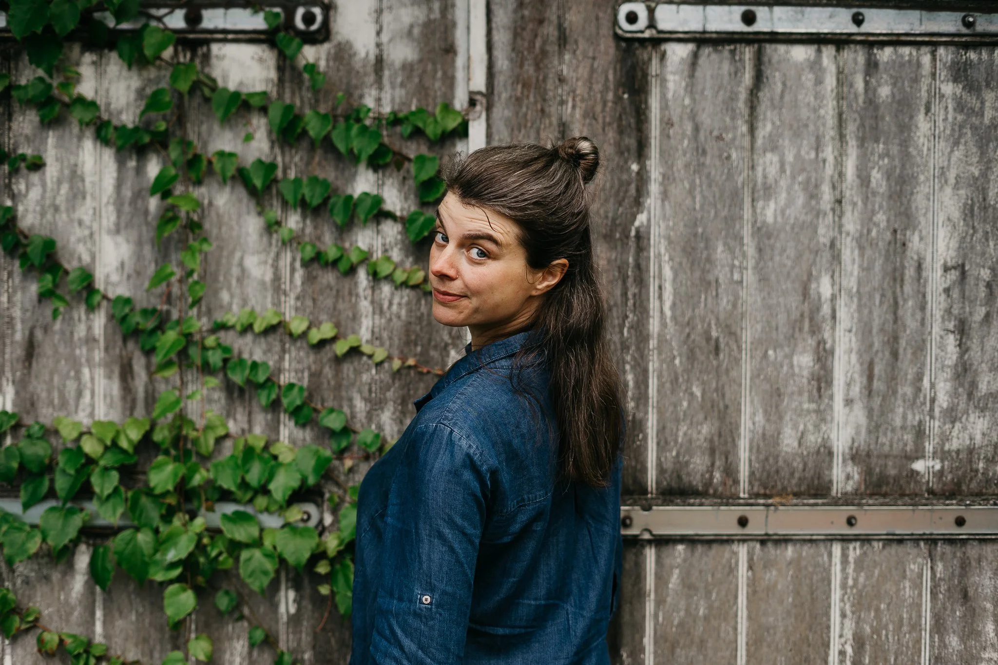 Een vrouw met donker haar in een knot, gekleed in een blauw jasje, staat naast een houten schutting met groene planten die langs de muur groeien.