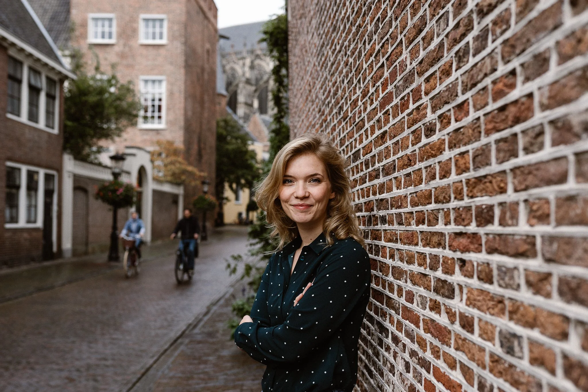 Vrouw met krullend haar, zwart met witte stippen blouse, staat met armen over elkaar naast een bakstenen muur, in een straat met oudere gebouwen en fietsers in de achtergrond.