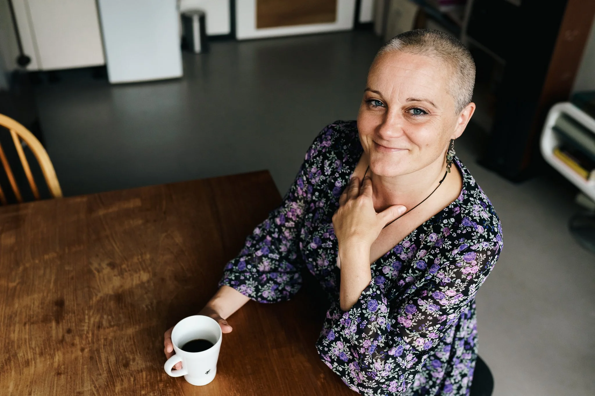 Een vrouw met een korte haarsnit en een bloemenjurk die een kop koffie vasthoudt en naar de camera glimlacht in een keuken.