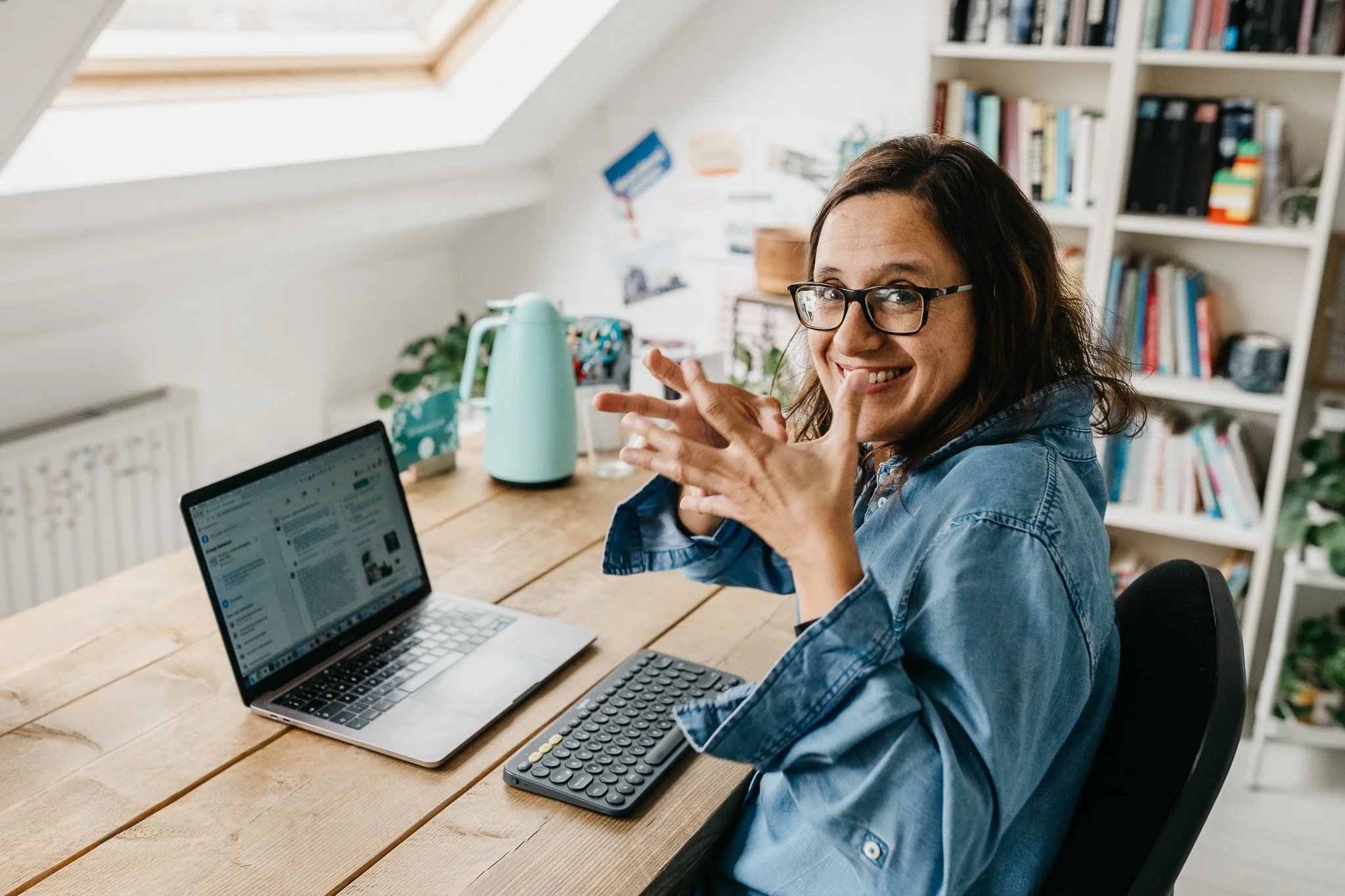 Vrouw zit aan een houten tafel in een lichte kamer, lachend en wijzend naar de camera, met een laptop, een computermuis, een toetsenbord, een waterkoker en boeken op de achtergrond.