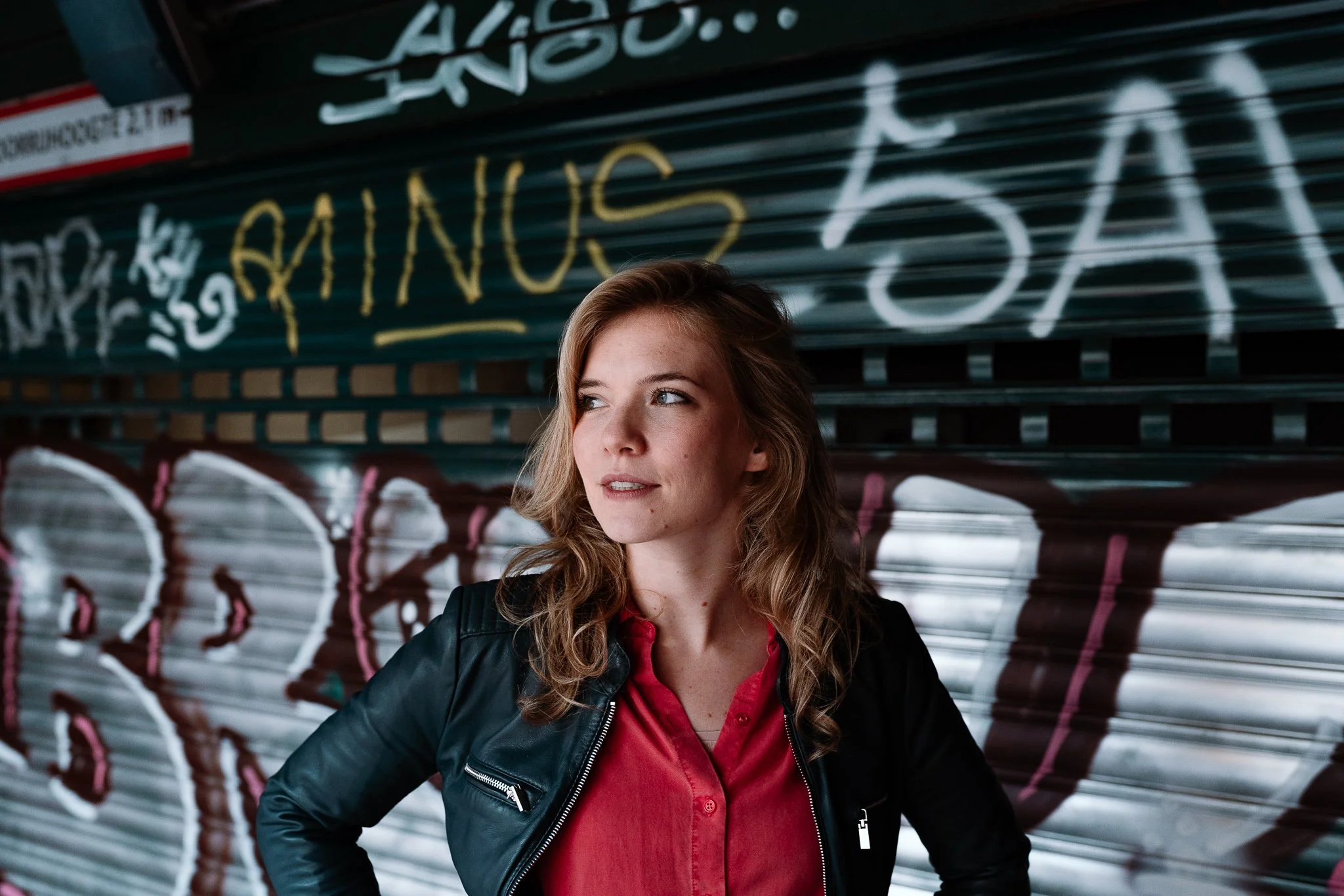 Een jonge vrouw met golvend haar staat voor een graffiti-bedekte muur en kijkt naar links.