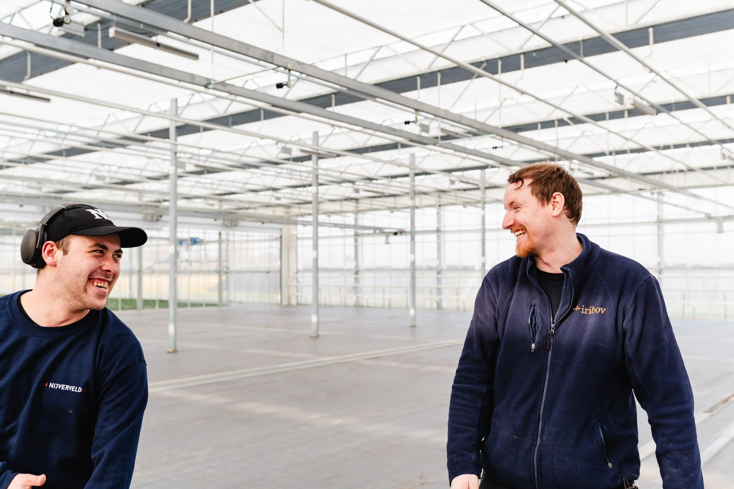 Two men laughing and talking inside a large, bright, empty greenhouse or industrial space with a metal framework and glass ceiling.