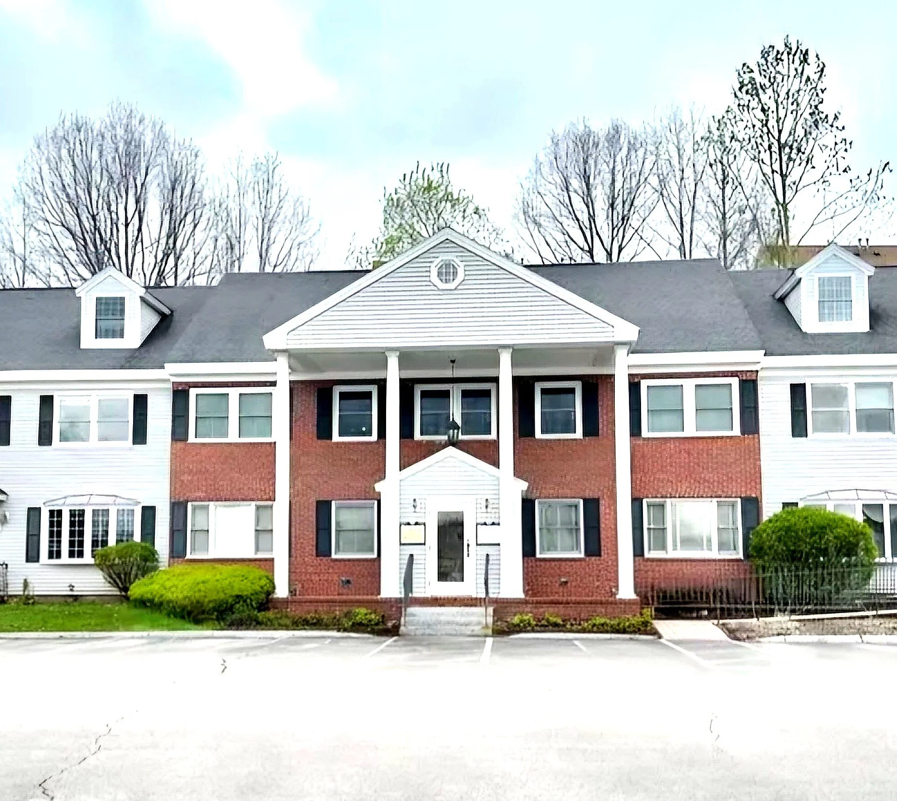 A three-story residential building with brick and white siding, a gray gabled roof, dormer windows, a central porch with white columns, surrounded by a green lawn, bushes, and trees in the background with a pale blue sky.