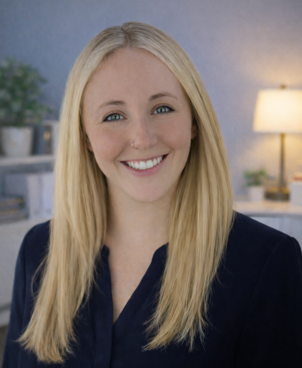 A smiling young woman with blonde hair, wearing a navy blue top, sitting in a cozy room with a lamp and plants in the background.