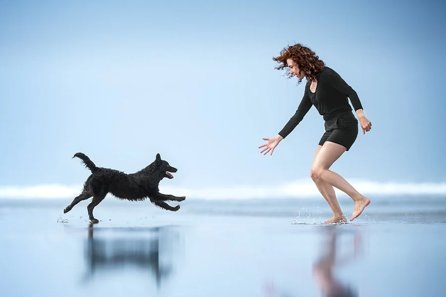 A black puppy runs towards a woman in black clothing at the beach during lowtide.