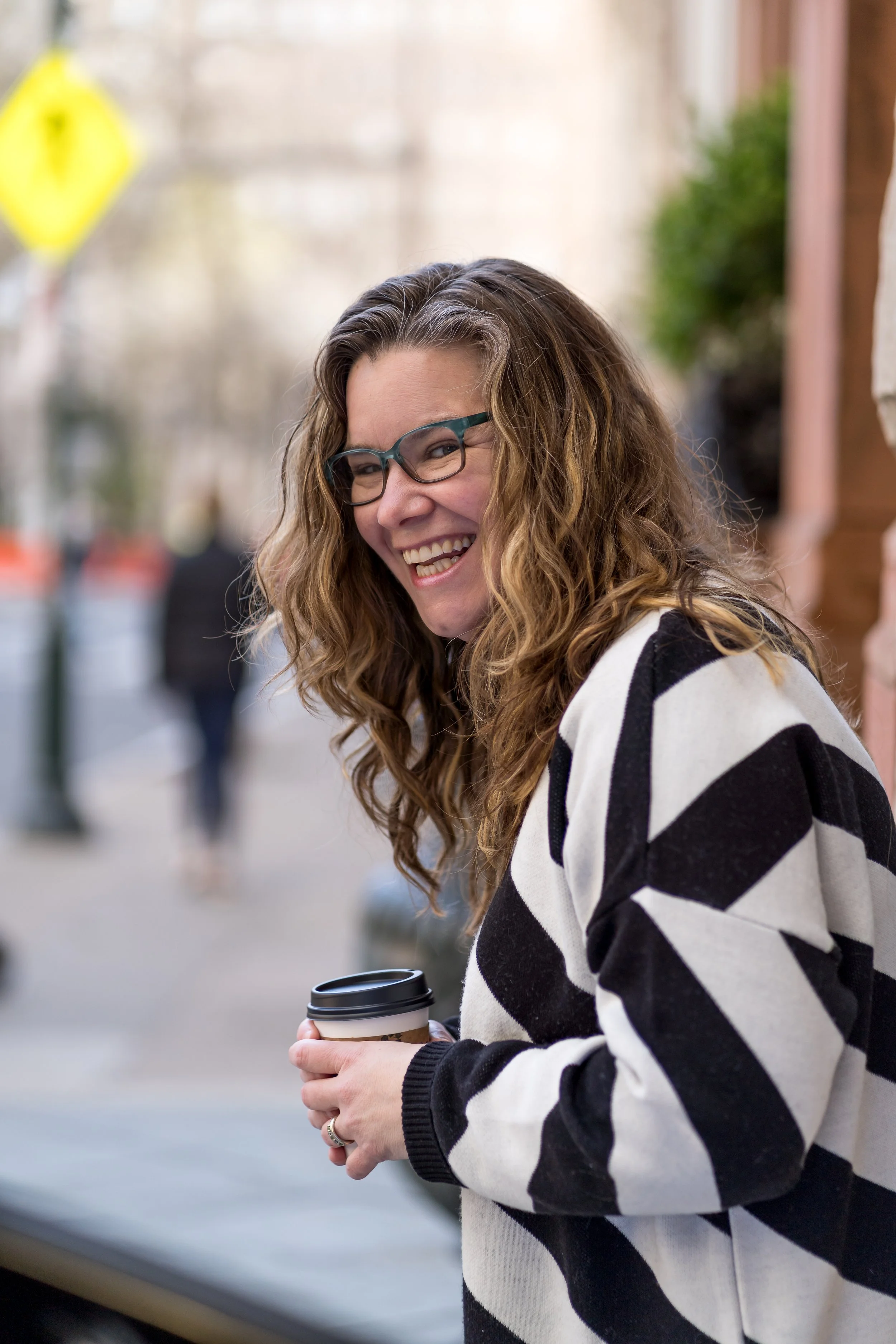 A woman with curly brown hair and glasses smiling while holding a coffee cup outdoors.