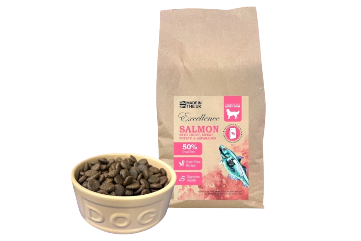 Dog food bag labeled 'Excellence Salmon with Trout, Sweet Potato & Asparagus' next to a white bowl filled with dry salmon dog food.