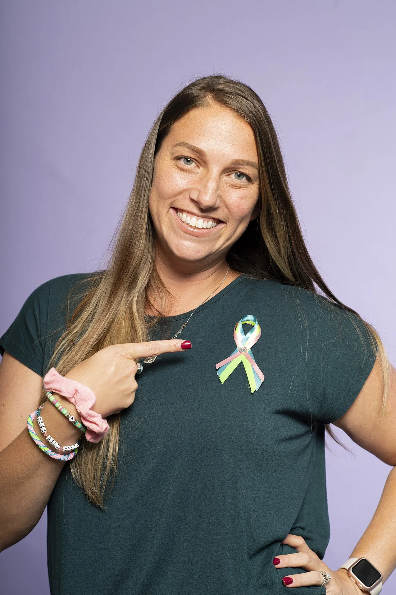 A woman with long brown hair smiling and pointing to a ribbon pin on her dark green shirt. She has a pink scrunchie, multiple bracelets, a ring, and a smartwatch. The background is purple.