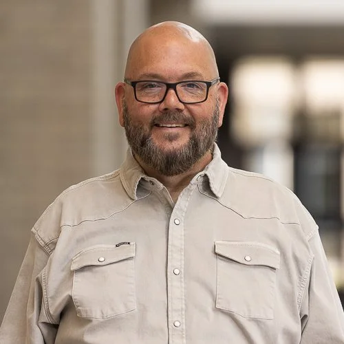 Smiling man with glasses and beard wearing a black hoodie with a logo, standing indoors.
