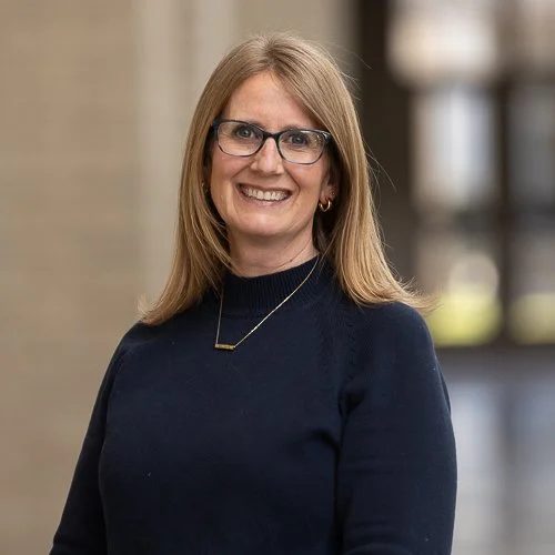 A smiling woman with shoulder-length blonde hair, wearing a white top, gold hoop earrings, and a small gold necklace, standing outdoors with a blurred urban background.