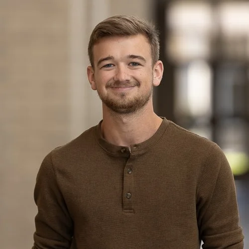 Young man with short brown hair, beard, and mustache, smiling, wearing a brown long-sleeve shirt, standing indoors with blurred background.