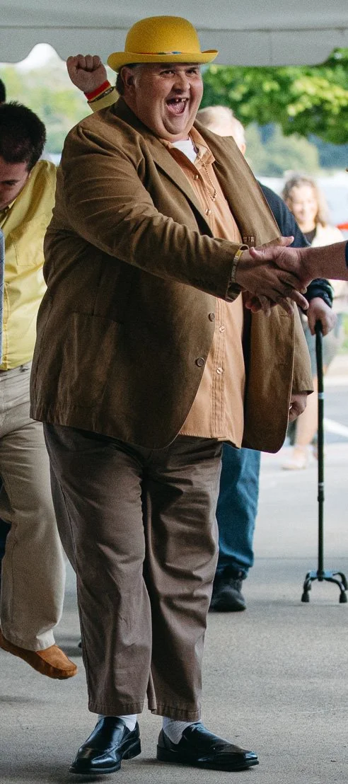 A person wearing a yellow hat and brown suit smiling and shaking hands at an outdoor event.