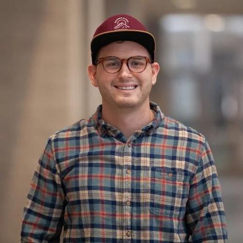 Smiling person wearing glasses, a plaid shirt, and a cap indoors