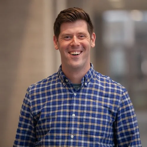 Smiling person wearing a blue plaid shirt in a blurred indoor setting.