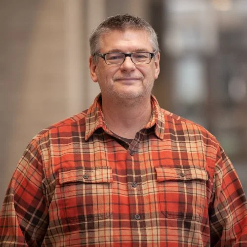 Smiling person wearing a red plaid shirt and glasses, standing indoors.