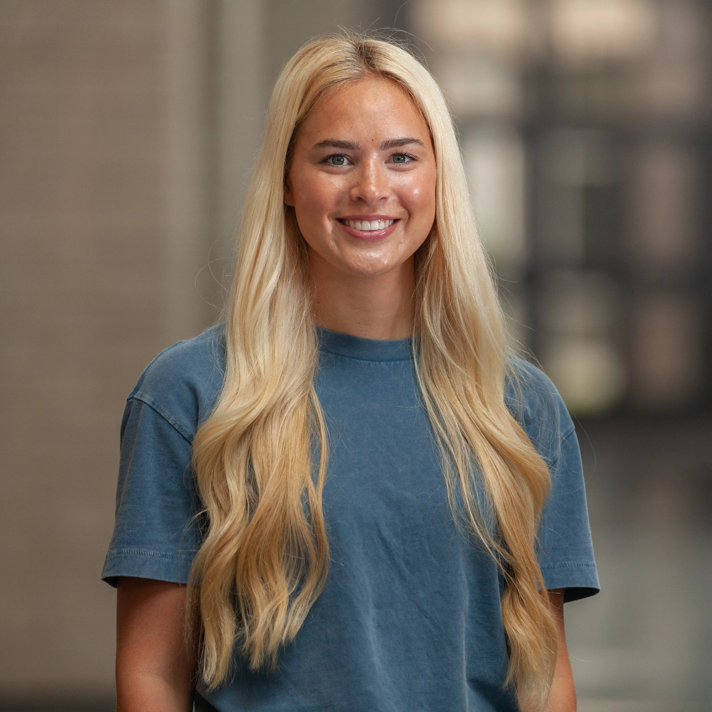 A woman with long blonde hair wearing a blue shirt, smiling, and standing indoors with a blurred background.