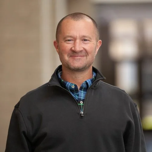 Man wearing a black zip-up sweater and plaid shirt, smiling in a blurred indoor setting.