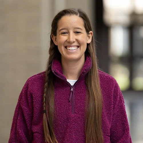 A woman with long brown hair wearing a maroon shirt smiles while standing outdoors.