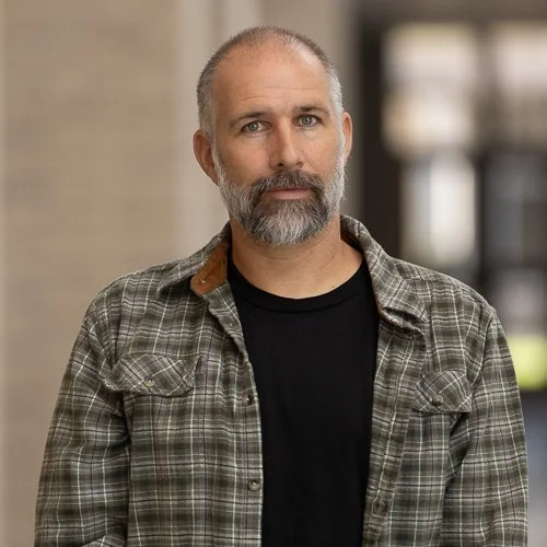 Man with a beard wearing a plaid shirt and black t-shirt standing indoors.