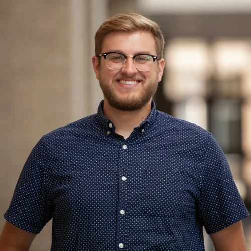 A person wearing a navy blue polka dot shirt and glasses, smiling in a well-lit indoor setting.
