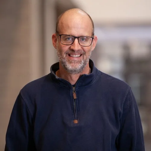 A smiling man wearing glasses and a navy blue pullover standing indoors.