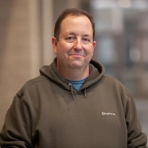 Person wearing a gray hoodie standing indoors with a blurred background.