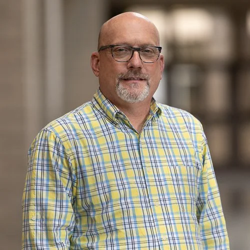 Man wearing glasses and a yellow plaid shirt standing indoors, blurred background.