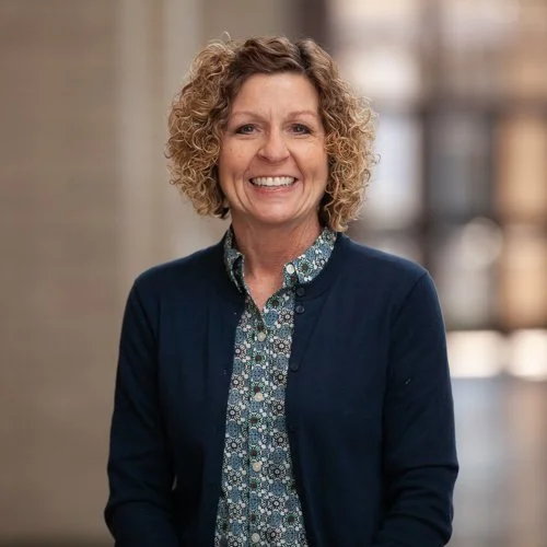 Person with curly hair smiling, wearing a dark cardigan and patterned shirt, standing indoors.