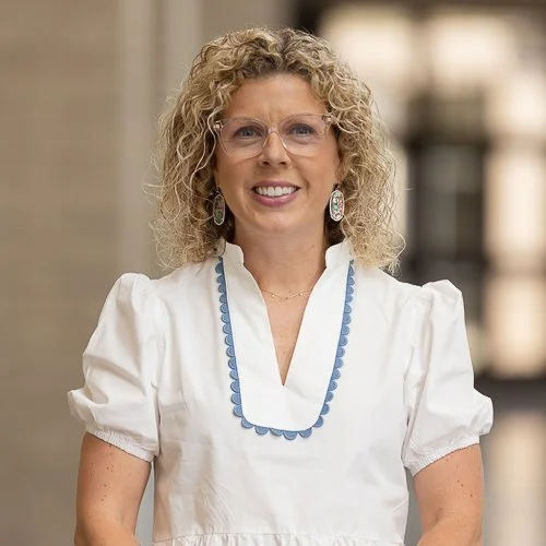 Smiling woman with curly blonde hair, wearing a leopard print blouse, standing indoors with a blurred background.