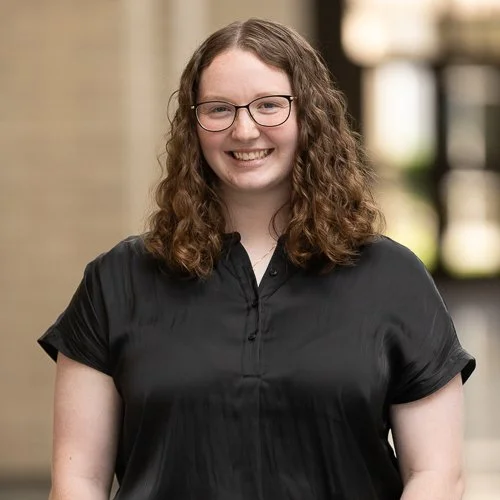 A smiling young woman with curly brown hair, wearing glasses and a black shirt, standing outdoors in front of a blurred background with trees and building structures.
