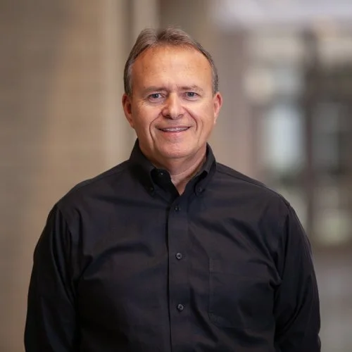 Man in black shirt standing indoors against a blurred background.