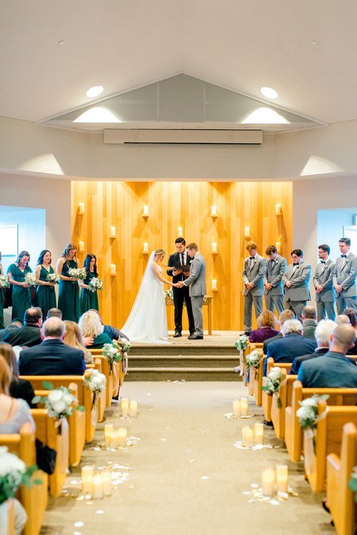 A wedding ceremony in a church with a couple exchanging vows, surrounded by bridesmaids in green dresses and groomsmen in gray suits. The aisle is decorated with flowers and candles, and guests are seated in pews.