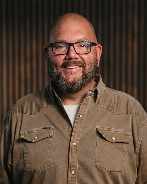 Smiling man with glasses and beard wearing a black hoodie with a logo, standing indoors.